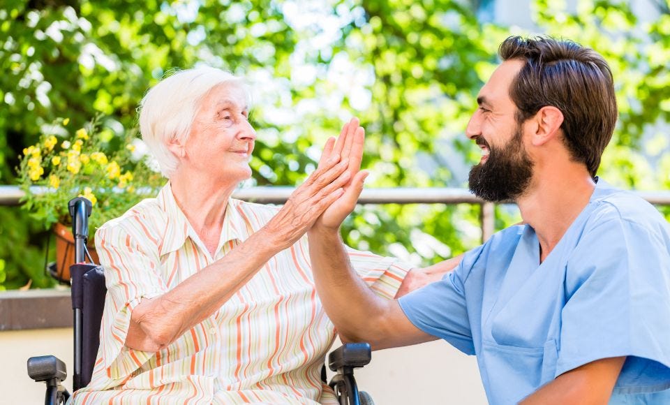 Ältere Dame im Rollstuhl gibt männlichem Pfleger die Hand.