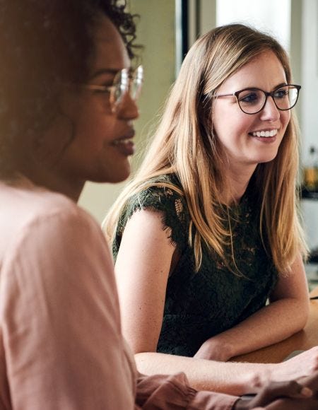 Zwei Frauen sitzen gemeinsam an einem Holztisch und arbeiten mit Laptop, Notizbuch und Kaffee in einem hellen Büro.