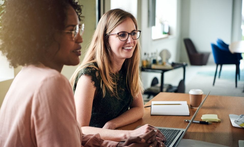 Zwei Frauen sitzen gemeinsam an einem Holztisch und arbeiten mit Laptop, Notizbuch und Kaffee in einem hellen Büro.