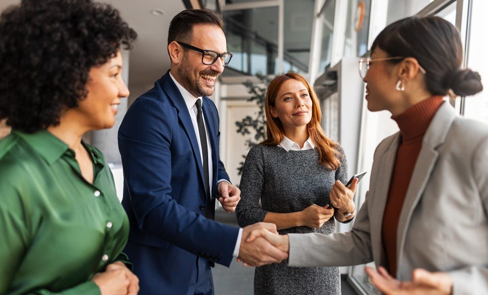 Mehrere Personen begrüßen sich per Handschlag in einem modernen Büro. Weitere Teilnehmende stehen daneben und verfolgen das Gespräch in professioneller Atmosphäre.