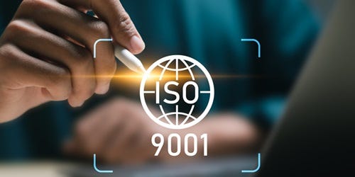 Person mit Stift, fokussiert auf ein leuchtendes ISO 9001-Logo vor einem verschwommenen Laptop.