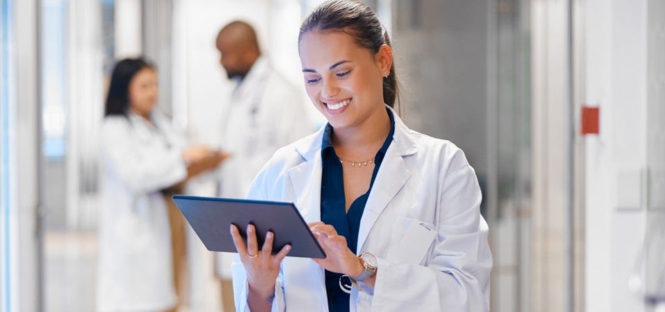 Qualitätsmanagementbeauftragte steht im Krankenhausflur und arbeitet lächelnd an einem Tablet, während Kollegen im Hintergrund sprechen.“