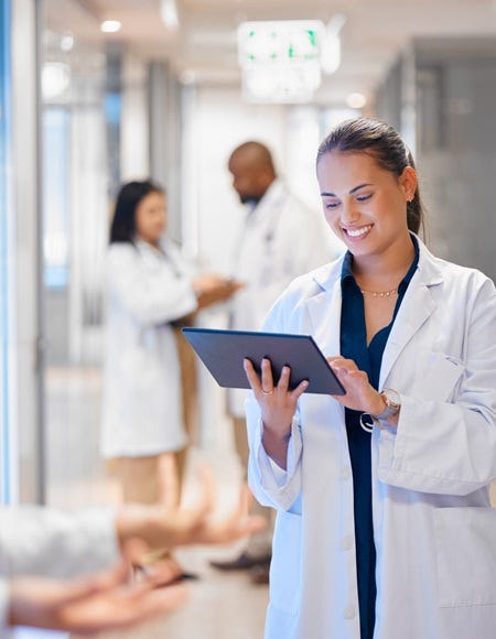 Qualitätsmanagementbeauftragte steht im Krankenhausflur und arbeitet lächelnd an einem Tablet, während Kollegen im Hintergrund sprechen.“