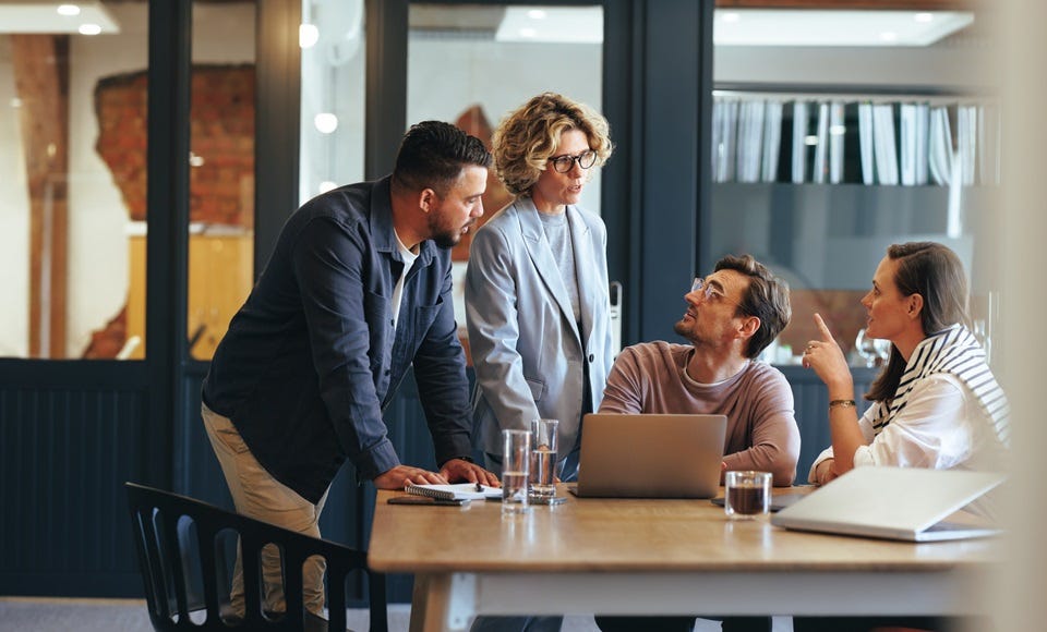 Mehrere Personen in einem modernen Büro bei einer Besprechung, mit Laptop, Notizbuch und Tablet auf einem Tisch, vor einer Backsteinwand.