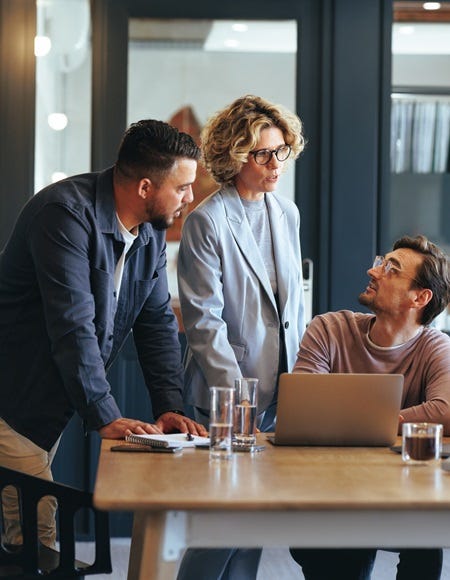 Mehrere Personen in einem modernen Büro bei einer Besprechung, mit Laptop, Notizbuch und Tablet auf einem Tisch, vor einer Backsteinwand.