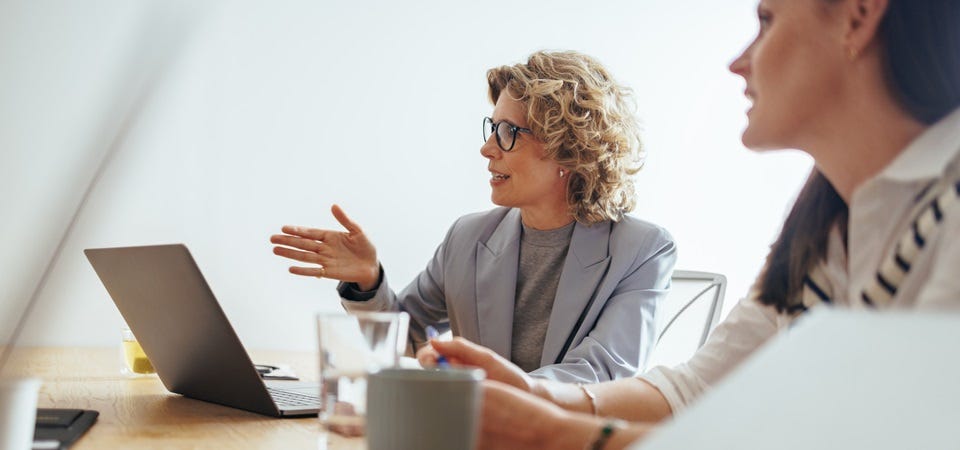 Weibliche F&uuml;hrungskraft spricht im Rahmen eines Meetings und unterstreicht das Gesagte mit Gesten. Auf dem Besprechungstisch stehen Laptop, Wasserglas und Kaffeetasse
