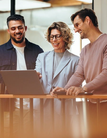 Drei Personen stehen zusammen an einem Gel&auml;nder und betrachten einen Laptop. Der Hintergrund zeigt ein modernes B&uuml;ro mit Holzelementen und gro&szlig;en Fenstern.