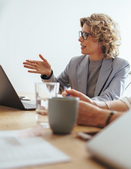 Weibliche F&uuml;hrungskraft spricht im Rahmen eines Meetings und unterstreicht das Gesagte mit Gesten. Auf dem Besprechungstisch stehen Laptop, Wasserglas und Kaffeetasse