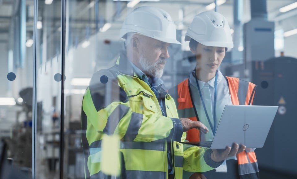 Zwei Personen mit Schutzhelmen und Warnwesten betrachten gemeinsam Energiekennzahlen auf einem Laptop in einer modernen Industriehalle.