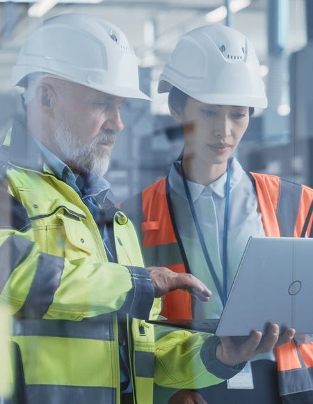 Zwei Personen mit Schutzhelmen und Warnwesten betrachten gemeinsam Energiekennzahlen auf einem Laptop in einer modernen Industriehalle.