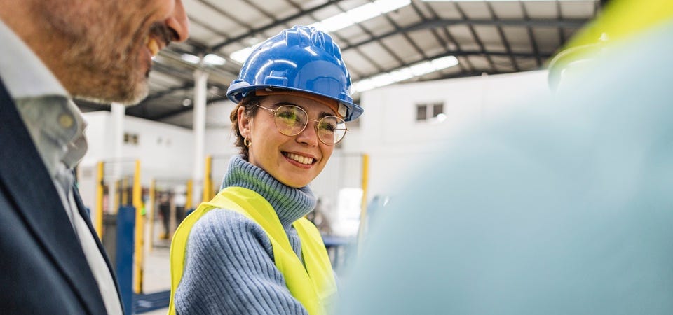Mitarbeiterin mit Schutzhelm und Warnweste im Gespr&auml;ch auf einem Industriegel&auml;nde.