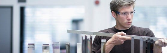 Das Bild zeigt einen Mann, der ein Metallstück betrachtet. Er trägt eine Schutzbrille und prüft, ob das Bauteil präzise gefertigt ist.