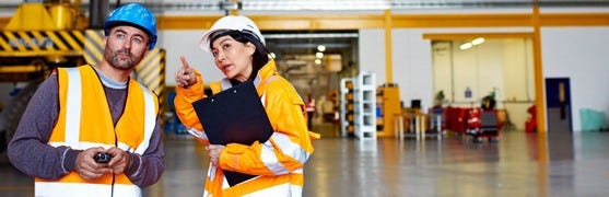 Ein Mann und eine Frau mit organefarbener Warnweste und Sichherheitshelm in einer Industriehalle, die sich gerade unterhalten