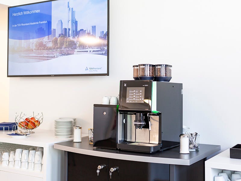 Schwarze Vollautomatkaffeemaschine, links daneben ein wei&szlig;er Schrank mit Kaffeebechern, &uuml;ber der Maschine h&auml;ngt an der wei&szlig;en Wand ein Flatscreen