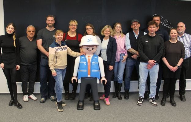13 Menschen stehen f&uuml;r ein Gruppenfoto zusammen. In der Mitte steht das Playmobil T&Uuml;V-M&auml;nnchen.