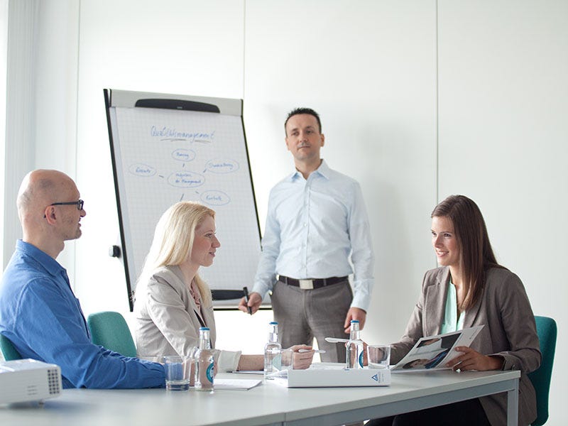 Schulungssituation: Zwei Frauen und ein Mann sitzen an einem Tisch und ein weiterer Mann steht an einem Whiteboard.