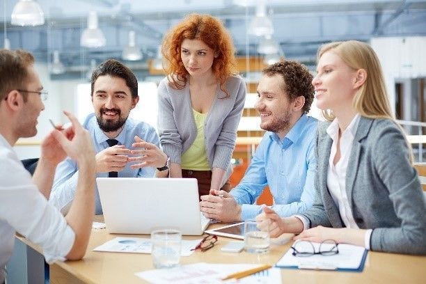 Fünf Menschen sitzen gemeinsam an einem Schreibtisch. Auf dem Tisch liegen Zettel, ein Laptop und Schreibmaterial. Sie unterhalten sich alle miteinander.