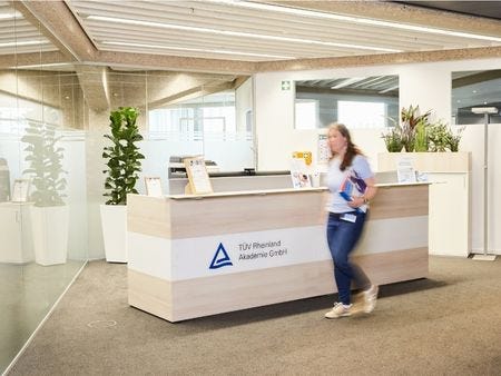 Empfangstresen TÜV Rheinland Akademie Hannover Eine Mitarbeiterin läuft am modernen Empfangstresen der TÜV Rheinland Akademie vorbei – der helle Holztresen mit Logo ist umgeben von Grünpflanzen und Informationsmaterialien in einem freundlichen Eingangsbereich.