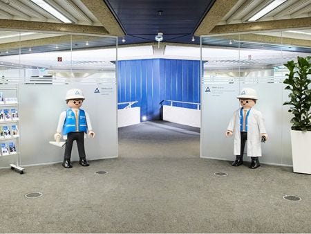 Eingangsbereich TÜV Rheinland Akademie Hannover Eingangsbereich mit zwei übergroßen Playmobil-Figuren – eine in Arbeitsschutzkleidung mit Helm und Warnweste, eine im weißen Laborkittel – die den Weg zum Durchgang flankieren.