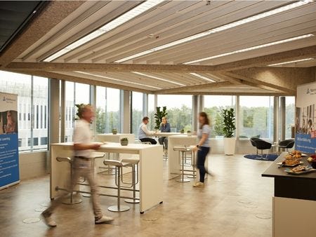 Bistrobereich Hannover Heller, offener Pausenbereich mit Stehtischen, Barstühlen und großen Fensterfronten – mehrere Personen bewegen sich durch den lichtdurchfluteten Raum mit Grünpflanzen und TÜV Rheinland Roll-ups im Hintergrund.