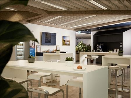 Bistrobereich TÜV Rheinland Akademie Hannover Moderne Lounge und Pausenbereich mit weißen Stehtischen, Barstühlen, Grünpflanzen, einem Bildschirm und einer Getränketheke – stimmungsvoll beleuchtet mit regionalem Bildmotiv an der Wand.