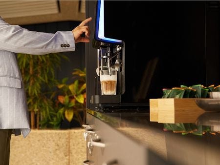 Kaffeemaschine im Bistrobereich Eine Person im hellen Blazer bedient eine moderne Kaffeemaschine im Bistrobereich - ein frisch zubereiteter Latte Macchiato steht bereit, im Hintergrund Grünpflanzen.
