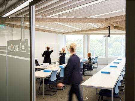 Seminarraum Kröpcke Blick an einerGlastür vorbei mit der Aufschrift 'Kröpcke' in einen Seminarraum – zwei Frauen arbeiten gemeinsam an einem Whiteboard, während Teilnehmende an langen Tischen mit blauen Ordnern sitzen und eine Person den Raum betritt.