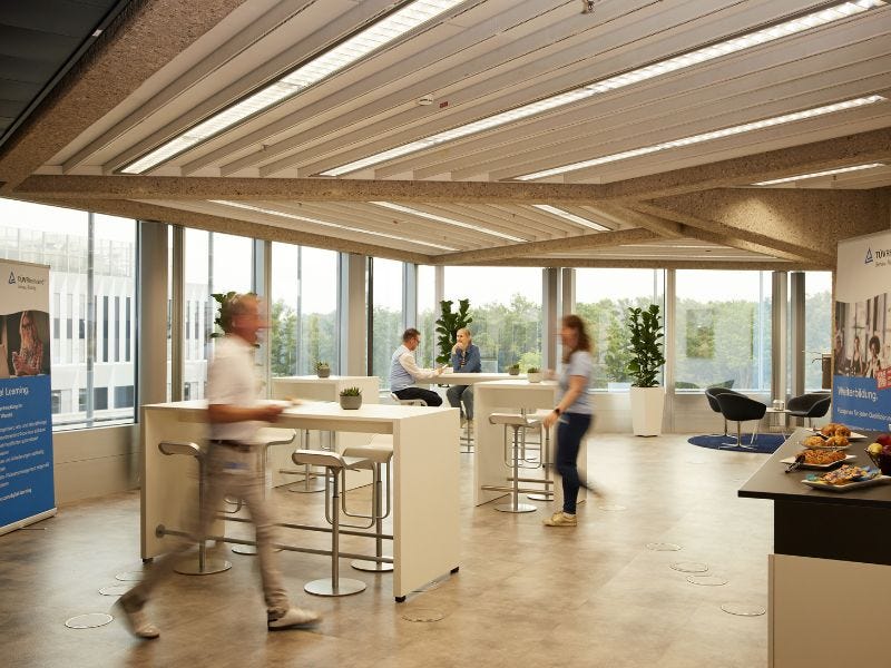 Das Bistro mit Steh- und Sitzgelegenheiten. Es gibt frisches Geb&auml;ck und Obst auf einem Buffet. Mehrere Menschen laufen durch den Raum. Durch die gro&szlig;en Fenster hat man einen Blick ins Gr&uuml;ne.