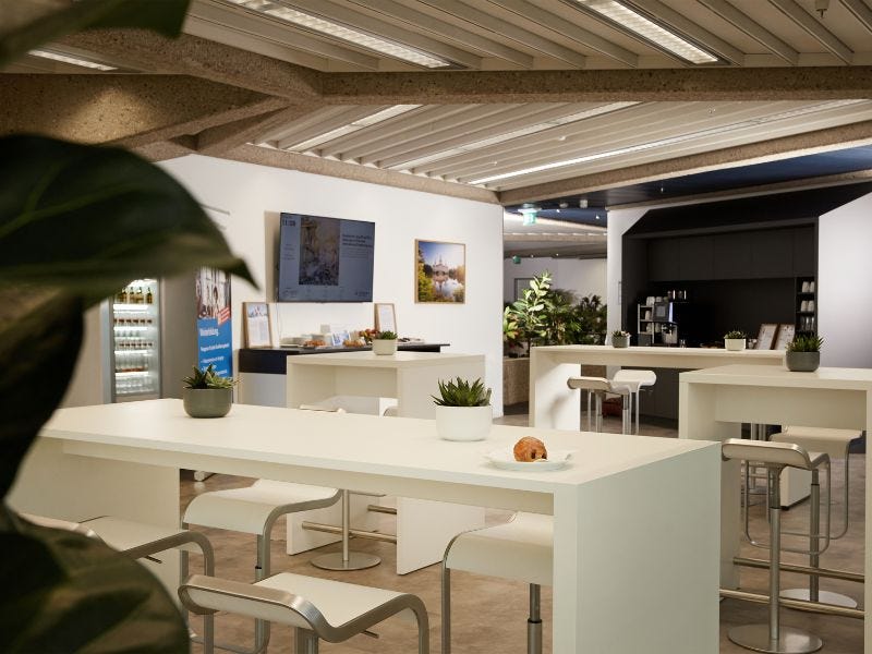 Bistrobereich TÜV Rheinland Akademie Hannover Moderne Lounge und Pausenbereich mit weißen Stehtischen, Barstühlen, Grünpflanzen, einem Bildschirm und einer Getränketheke – stimmungsvoll beleuchtet mit regionalem Bildmotiv an der Wand.