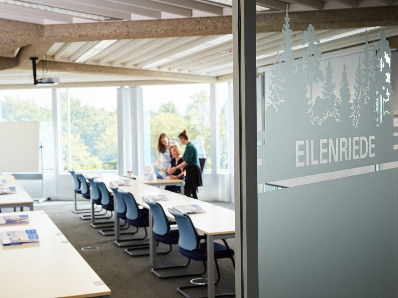 Blick in einen hellen Seminarraum mit modernen St&uuml;hlen. Im Hintergrund sind Fenster mit Blick ins Gr&uuml;ne. Auf der Glast&uuml;r steht der Raumname "Eilenriede".