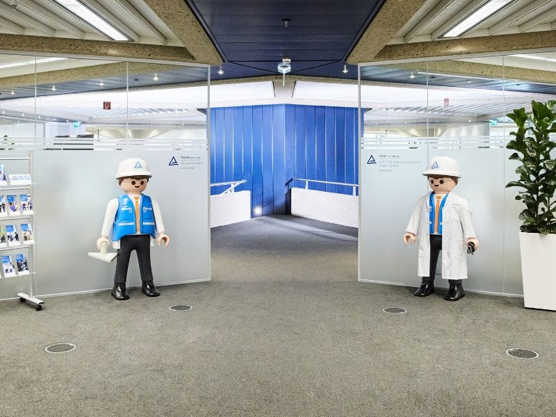 Eingangsbereich TÜV Rheinland Akademie Hannover Eingangsbereich mit zwei übergroßen Playmobil-Figuren – eine in Arbeitsschutzkleidung mit Helm und Warnweste, eine im weißen Laborkittel – die den Weg zum Durchgang flankieren.