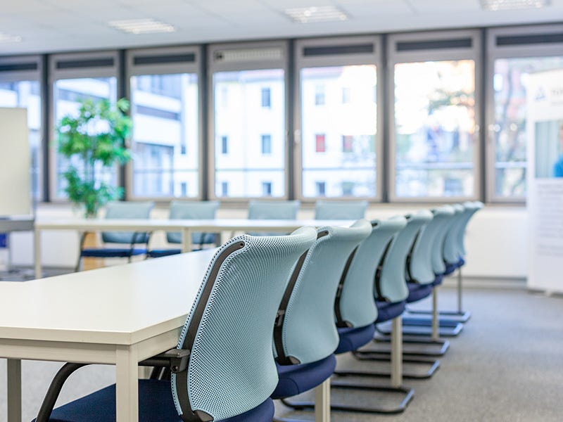 Ein heller Seminarraum mit modernen, ergonomischen Stühlen in einer Reihe entlang eines weißen Konferenztisches. Im Hintergrund sind Fenster mit Blick auf ein benachbartes Gebäude, ein Flipchart und eine Zimmerpflanze zu sehen. Der Raum wirkt freundlich.