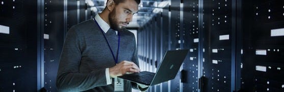 Das Bild zeigt eine Person, die in einem modernen Serverraum arbeitet. Die Person ist mit einem grauen Pullover und einem weißen Hemd bekleidet und sitzt vor einem Laptop. Im Hintergrund sieht man mehrere Reihen von Hochleistungs-Servern.