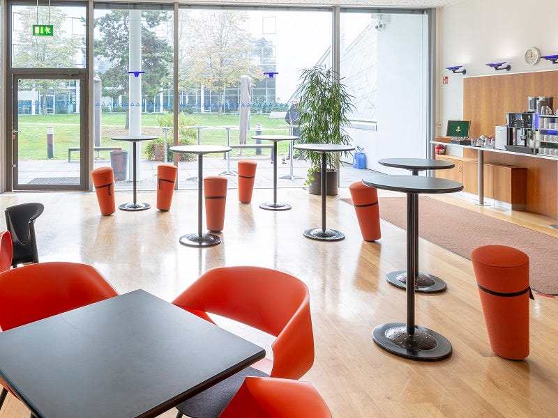 Heller Lounge-Bereich mit Stehtischen, orangen Hockern, Tischen, Sesseln, Kaffeestation und Fensterfront mit Gartenblick.