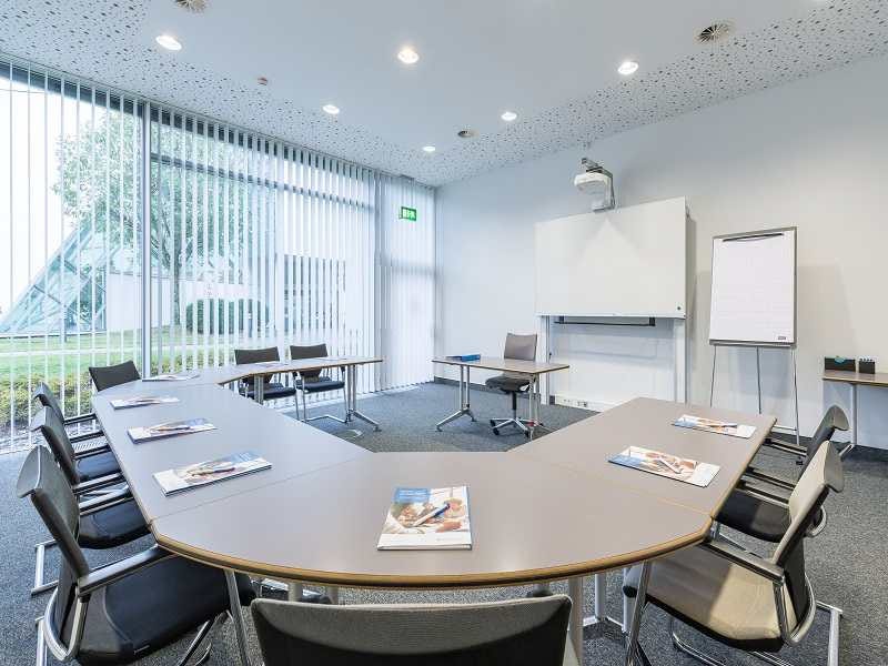 Heller Tagungsraum mit U-f&ouml;rmiger Tischanordnung, schwarzen St&uuml;hlen, Whiteboard, Flipchart und vertikalen Fensterjalousien.
