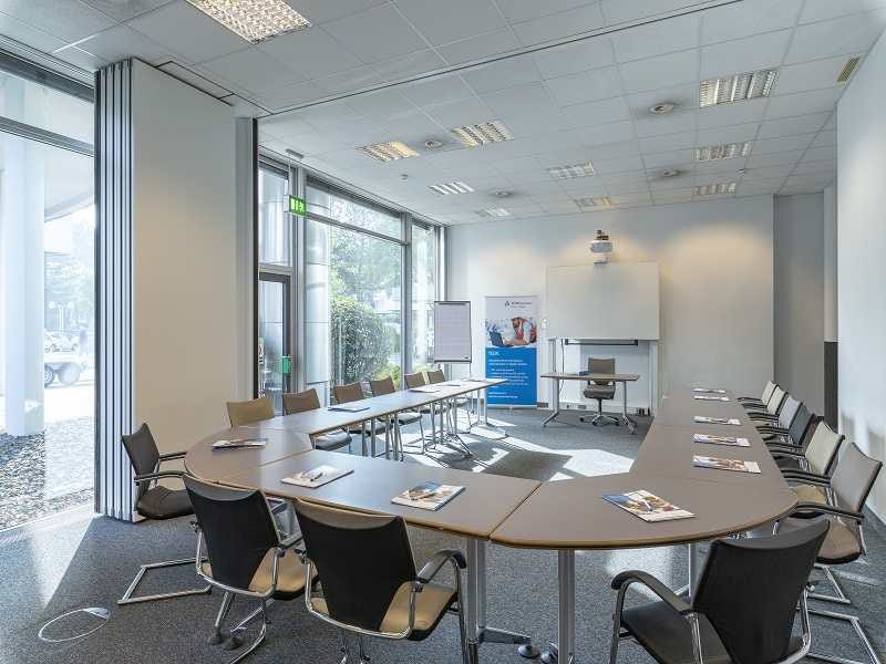 Heller Tagungsraum mit U-f&ouml;rmiger Tischanordnung, schwarzen St&uuml;hlen, Flipchart und Banner vor gro&szlig;en Fensterfronten.