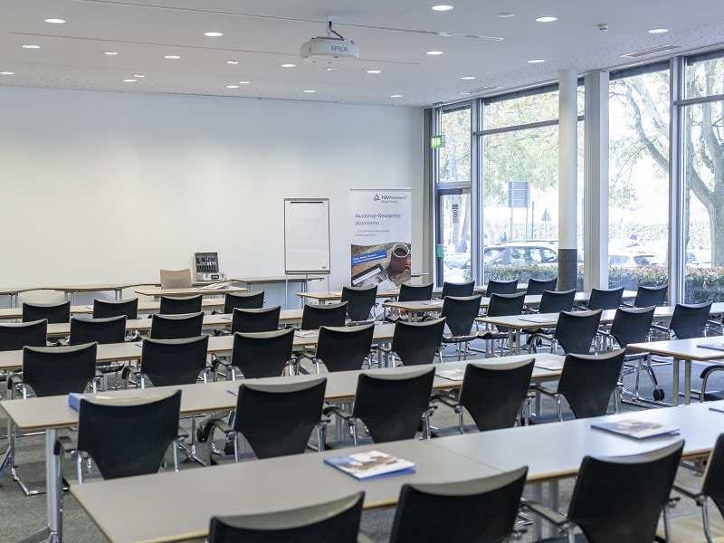 Lichtdurchfluteter Seminarraum mit Reihenarbeitspl&auml;tzen, schwarzen St&uuml;hlen, Deckenprojektor und Banner vor gro&szlig;en Fenstern.