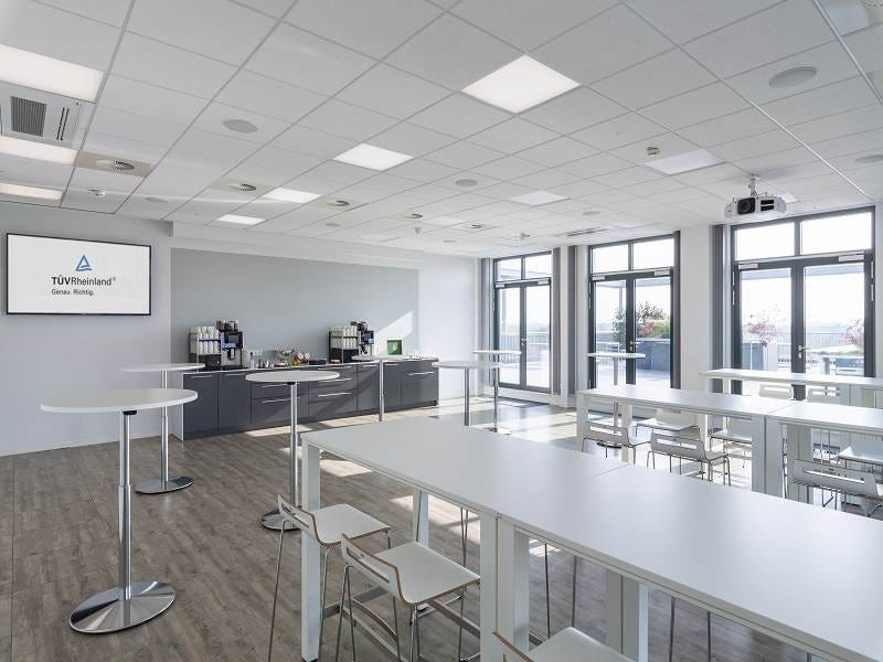 Heller Lounge-Bereich mit hohen Tischen, Barhockern, Kaffee-Station, Wanddisplay mit T&Uuml;V Rheinland-Logo und Terrassenblick.