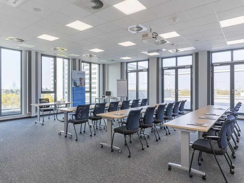 Heller Seminarraum mit zwei Reihen von Tischen, blauen St&uuml;hlen, Deckenprojektor, Banner und Flipchart vor bodentiefen Fenstern.