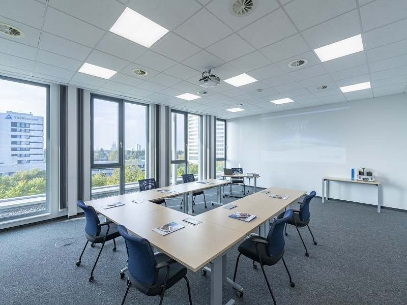 Heller Tagungsraum mit U-f&ouml;rmig angeordneten Tischen, blauen Drehst&uuml;hlen, Deckenprojektor und gro&szlig;er Fensterfront mit Blick auf umliegende Geb&auml;ude.