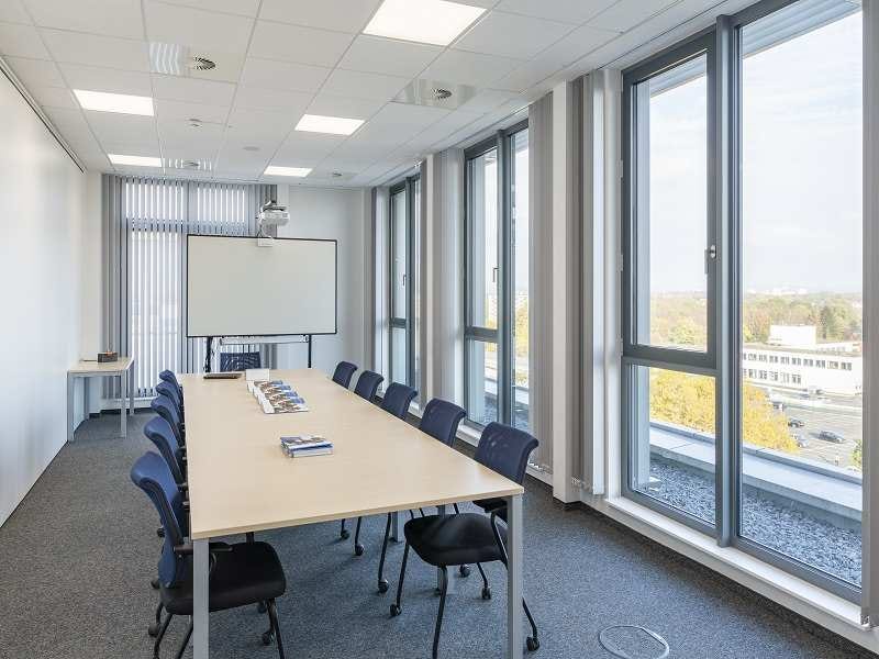 Heller Konferenzraum mit langem Tisch, schwarzen St&uuml;hlen, mobiler Leinwand, Deckenprojektor und bodentiefen Fenstern mit Jalousien.