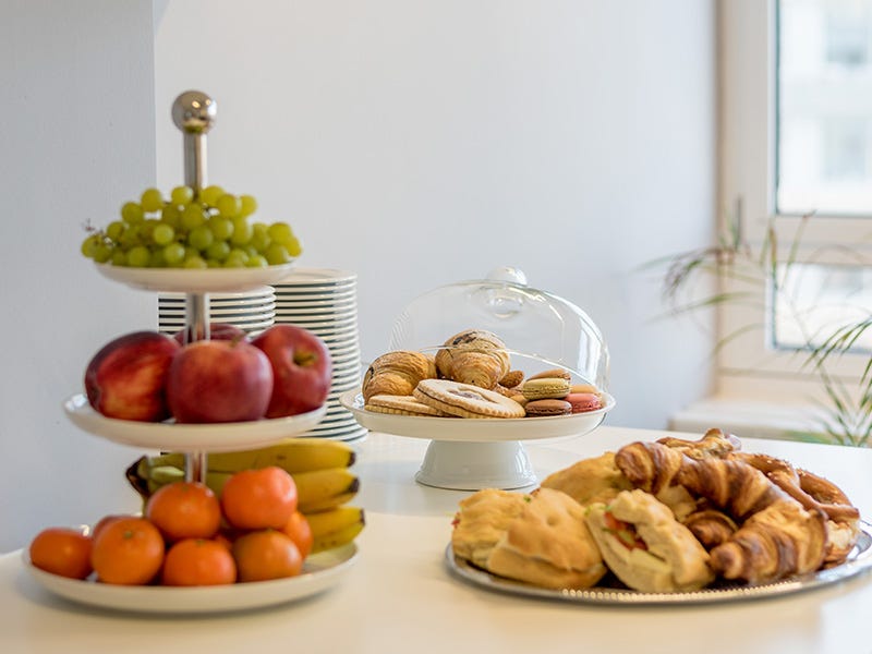 Das Bild zeigt einen Tisch mit einer Auswahl an frischem Obst und Gebäck. Auf einem dreistöckigen Tellerständer sind Äpfel, Mandarinen, Bananen und Trauben angerichtet. Daneben stehen auf Tellern verschiedene Backwaren wie Croissants und Brötchen bereit.