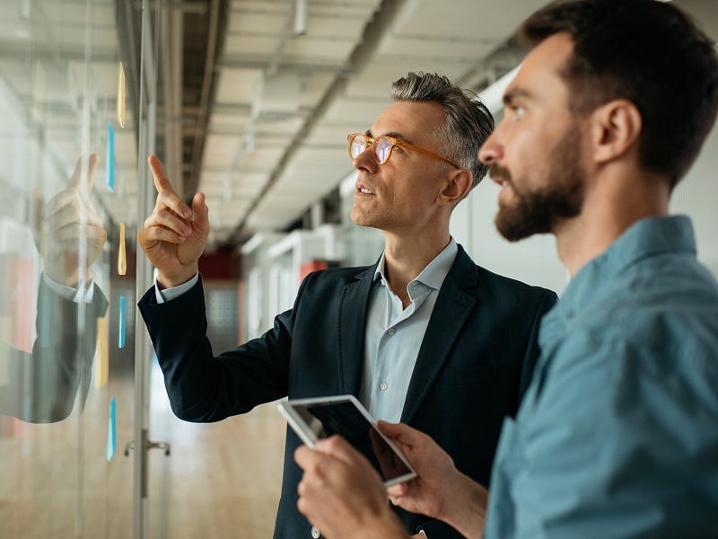 Das Bild zeigt zwei Personen in einem Geschäftsumfeld. Eine Person, die in einem Anzug gekleidet ist, gestikuliert und erklärt etwas, während die andere Person mit einem Tablet in der Hand zuhört. Im Hintergrund ist eine moderne Büroumgebung zu sehen.