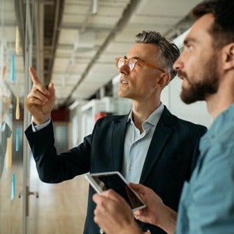 Das Bild zeigt zwei Personen in einem Geschäftsumfeld. Eine Person, die in einem Anzug gekleidet ist, gestikuliert und erklärt etwas, während die andere Person mit einem Tablet in der Hand zuhört. Im Hintergrund ist eine moderne Büroumgebung zu sehen.
