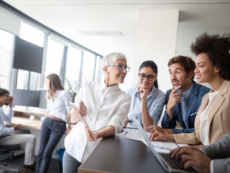Mehrere Berufstätige in einem modernen Büro tauschen sich in lockerer Atmosphäre aus, während eine Frau mit Tablet etwas präsentiert.