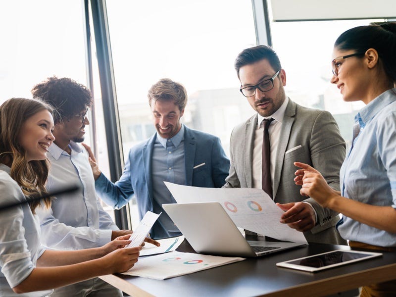 Das Bild zeigt eine Gruppe von Prsonen, die sich in einem modernen Büro an einem Tisch versammelt haben. Sie diskutieren angeregt über Dokumente und nutzen einen Laptop, um Informationen zu teilen. Große