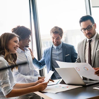 Das Bild zeigt eine Gruppe von Prsonen, die sich in einem modernen Büro an einem Tisch versammelt haben. Sie diskutieren angeregt über Dokumente und nutzen einen Laptop, um Informationen zu teilen. Große