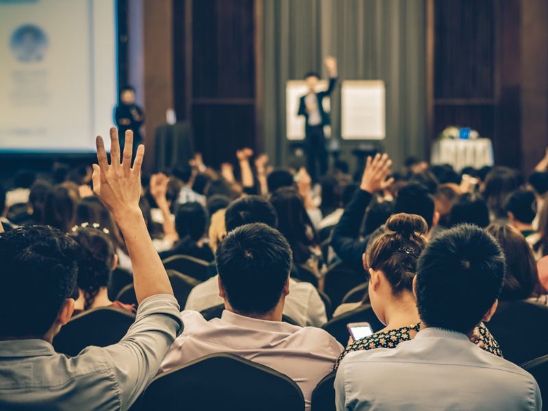 Publikum bei einer Konferenz oder Schulung – Teilnehmende sitzen in Reihen, einige heben die Hand, im Hintergrund spricht ein Moderator vor einer Präsentation.