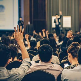 Publikum bei einer Konferenz oder Schulung – Teilnehmende sitzen in Reihen, einige heben die Hand, im Hintergrund spricht ein Moderator vor einer Präsentation.