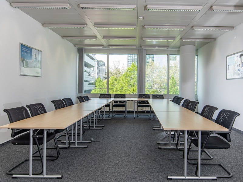 Das Bild zeigt einen modernen Seminarraum mit langen Tischen und schwarzen Stühlen. Große Fenster sorgen für viel natürliches Licht und bieten einen Blick ins Grüne. Die Raumgestaltung ist funktional und schlicht.